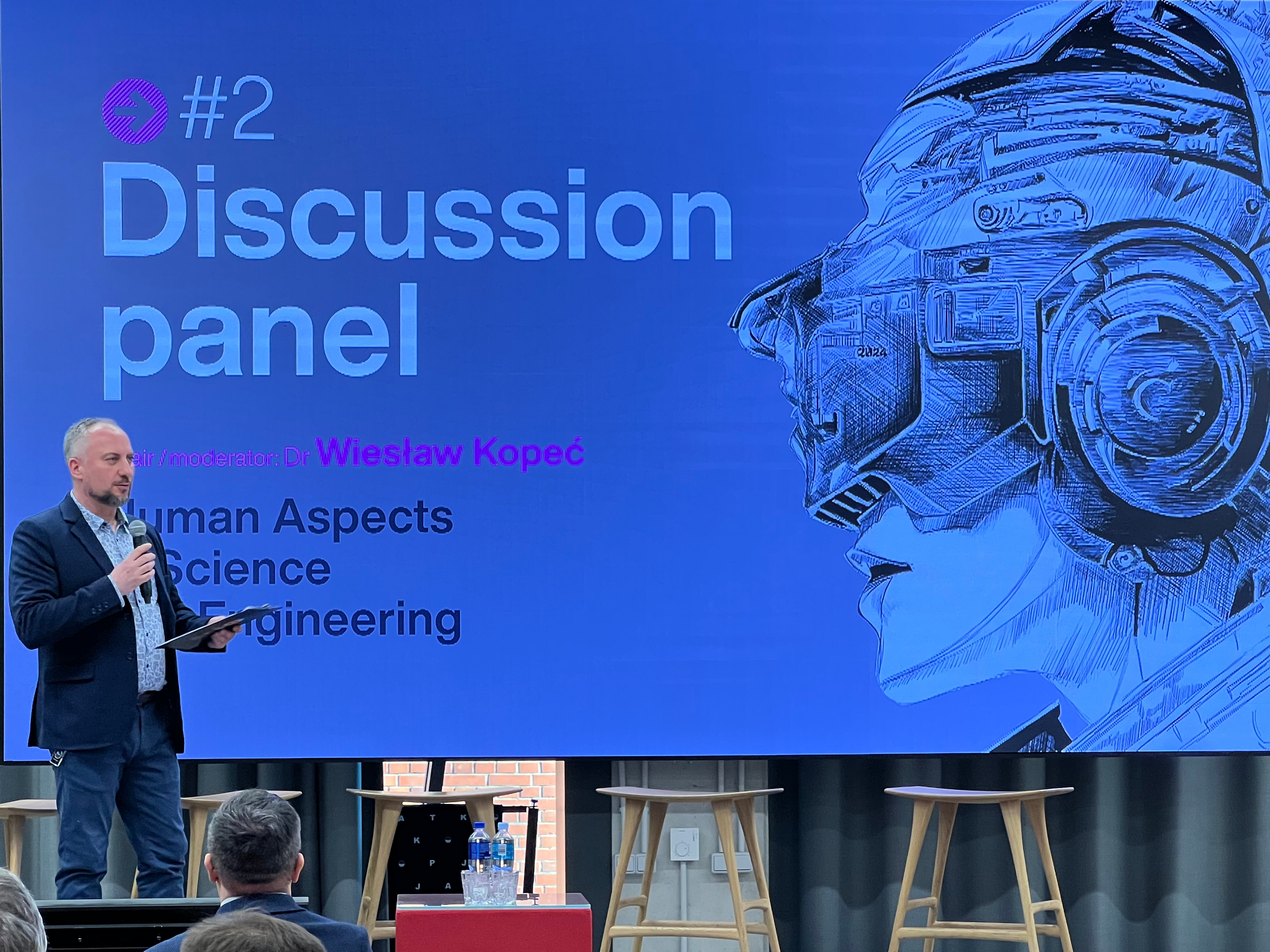 A photo of the conference's chair announcing a panel discussion. In the background a huge screen with logo of the conference, and panel information